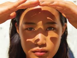 Woman shielding eyes from sunlight with hands, strong shadow on face, outdoor setting