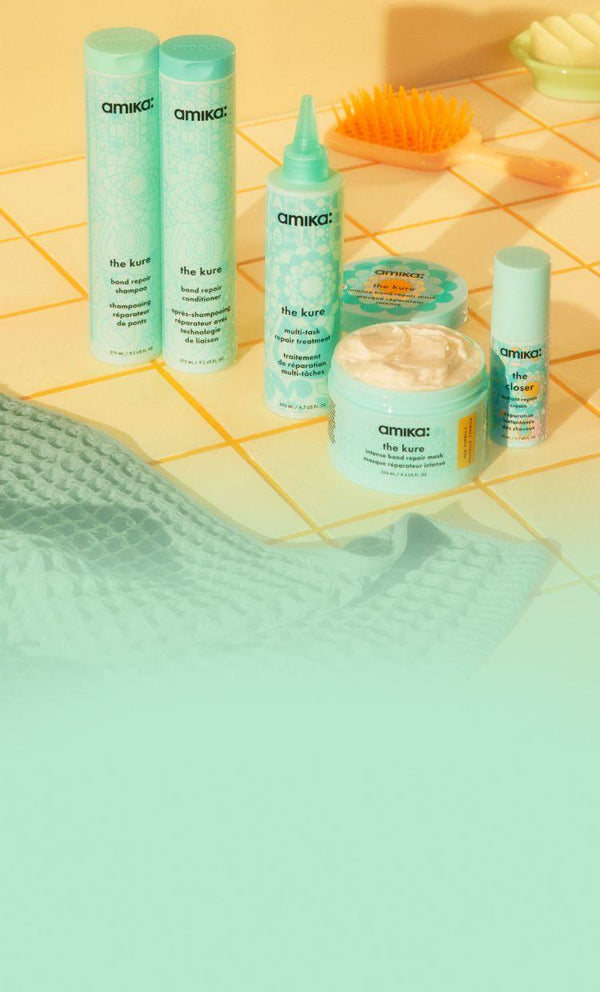 Amika The Kure shampoo, conditioner, and hair mask products on yellow tiled bathroom counter.